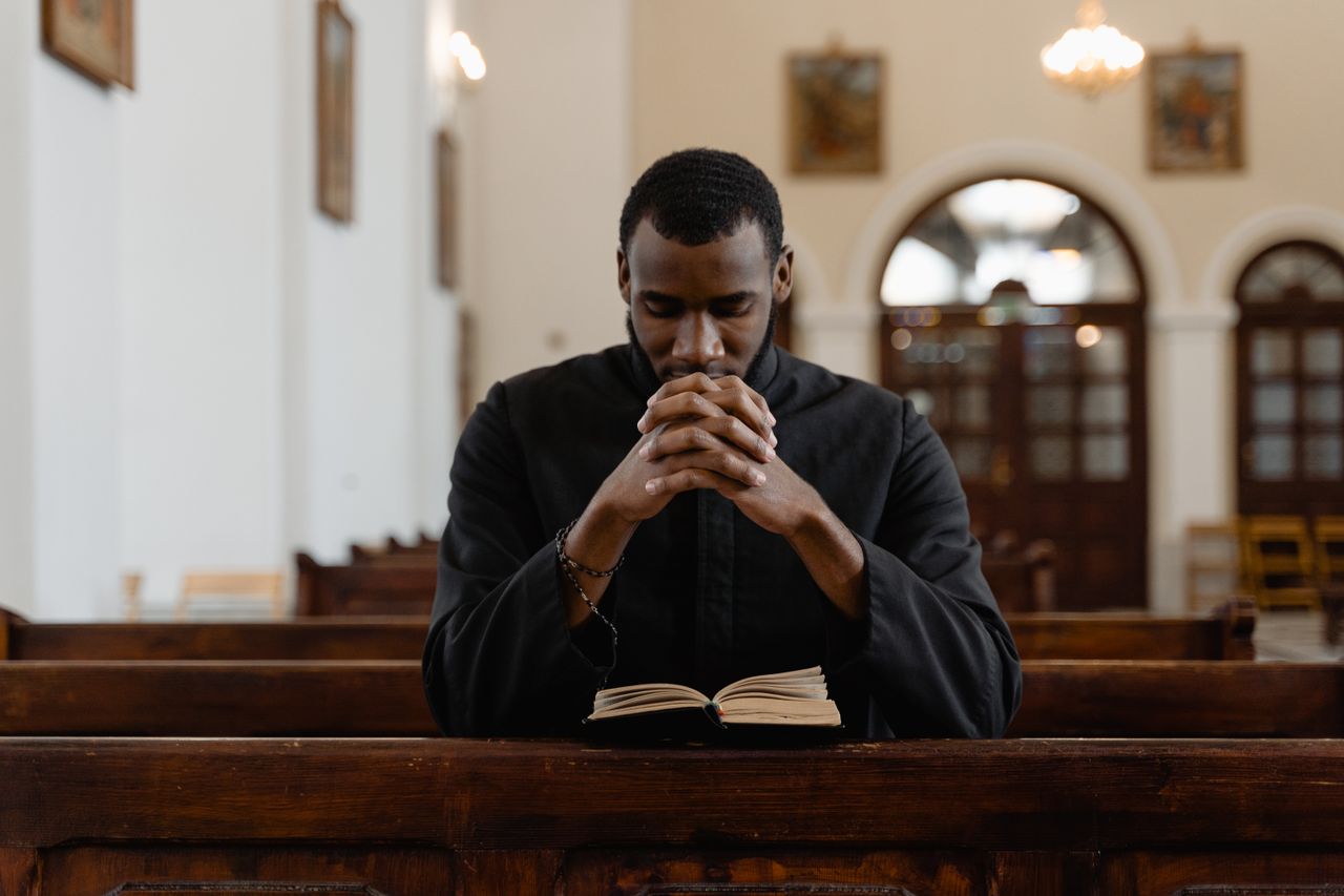 A man in religious attire prays in a church pew with an open Bible, reflecting faith.