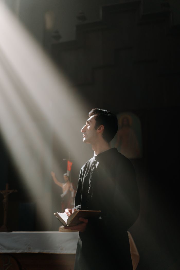 A priest stands in a sunlit church, holding a Bible with reverent expression.